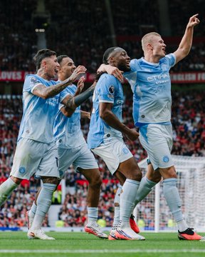 Four Manchester City football players in light blue jerseys celebrating together on the pitch, with one player pointing upward, in front of a stadium crowd.