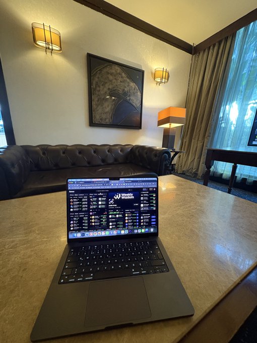 A photograph of a laptop on a large wooden table in an elegant room with a leather Chesterfield sofa, wall sconces, a framed artwork, and curtains, with the laptop screen displaying a cryptocurrency exchange interface with multiple trading pairs.