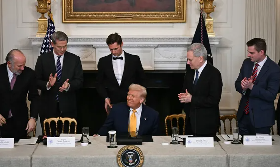 Donald Trump sits at a table in the White House surrounded by men in suits who are applauding; visible name cards read 'David Bessler' and 'Patrick Hruby'.