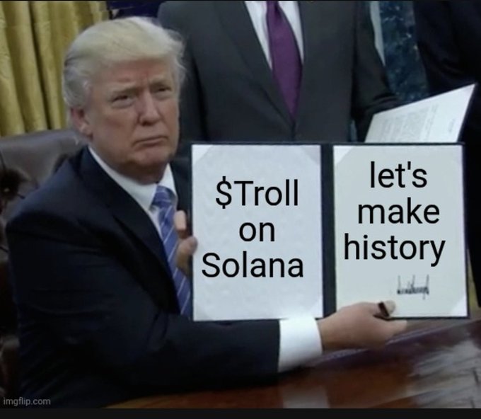 In the image, a man sits at a desk, sporting a serious demeanor and dressed in a dark suit with a red and blue tie. He holds two signs reading "$Troll" and "Let's make history", alluding to the ticker symbol "$TROLL" mentioned in the accompanying post. His seat is positioned in front of a well-appointed office backdrop.