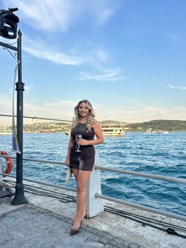 The image features a woman standing near a railing with her hand on her hip, holding a glass of wine. She appears to be posing for a photo while enjoying her drink. The background shows a beautiful view of the Bosphorus strait or bay, which adds an interesting and picturesque element to the scene. The post includes text that reads "Beautiful chats about the future of Web3, accompanied by a Bosphorus view. We're at the Stellar event today." This suggests that the woman is attending a social gathering or event related to cryptocurrency or web3 technology.