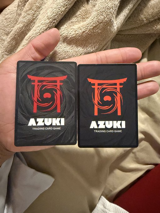 A hand holds two "Azuki Trading Card Game" cards for comparison—one featuring a swirling black-and-gray design (alpha edition) and the other a simpler dotted pattern (1st set edition)—showcasing why the alpha set's more detailed backside is often considered the more striking design.