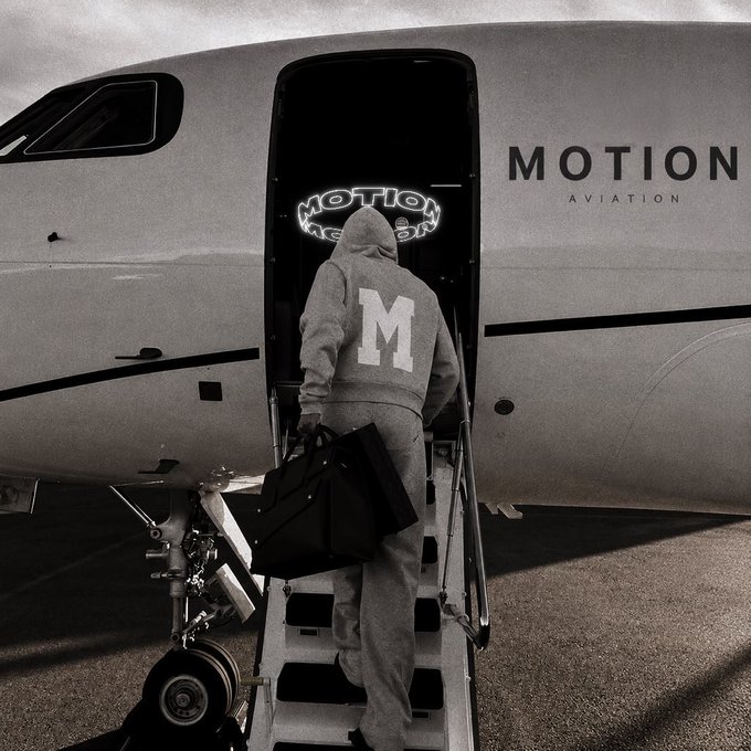 A black and white photo of a person in a hoodie with a large 'M' on the back boarding a 'MOTION AVIATION' private jet, carrying a black bag.