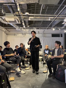 A man in dark clothing speaking into a microphone at what appears to be an indoor event or workshop, with seated attendees visible in the background.