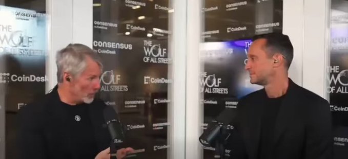 Two men in dark clothing hold microphones during an interview at the Consensus by CoinDesk event, with "THE WOLF OF ALL STREETS" branding visible on the backdrop.