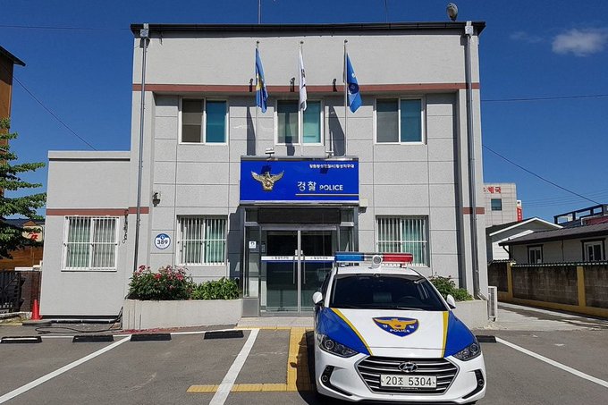 The image shows a white van with blue accents, possibly a police vehicle or a patrol car, parked in front of a large building. The building appears to be a government or police office. Above the van, there are three flags fluttering - one representing Korean culture and the other two representing Japan and Russia. In addition, there is text on the image that reads: "INTEL: South Korean police arrest 23 people in ร้านยงสมิดพฒครียกทรเฟลม", indicating a case related to the laundering of Nazi uniforms, with intelligence services being involved.