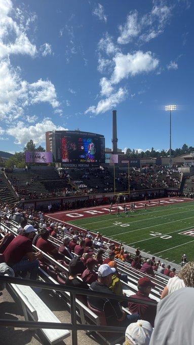 A view from the stands of a football stadium where fans watch a game on a sunny day, with 'AGGIES' painted in the end zone and a large screen displaying game footage.