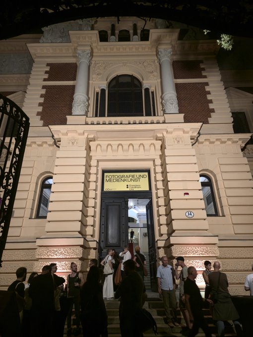 Nighttime exterior of an ornate historic building illuminated in warm light, with a crowd gathered outside. The entrance sign reads “GEOTGRAF? UND MEDIENKUNST” (partially stylized), and a small plaque marked “14” is visible to the right.