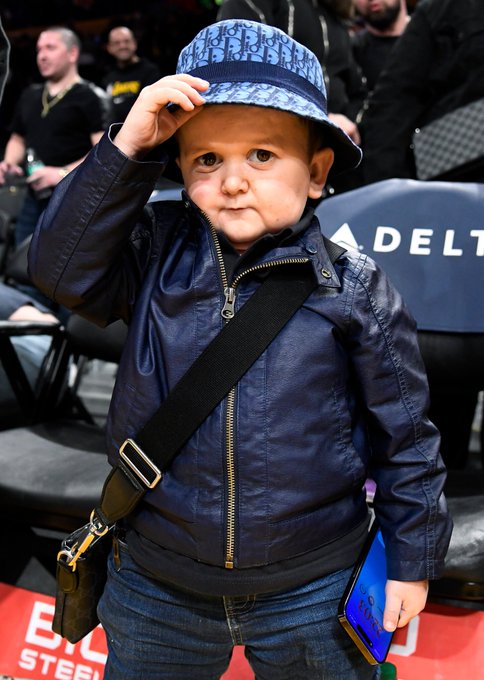 A young child stands at an event wearing a navy blue jacket with a crossbody bag and a blue Christian Dior bucket hat, with 'DELTA' signage visible in the background.