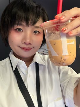 A selfie of a young Asian woman with short dark hair wearing a white collared shirt, holding up an iced bubble tea drink with a red straw in a clear cup branded with 'TE' lettering.