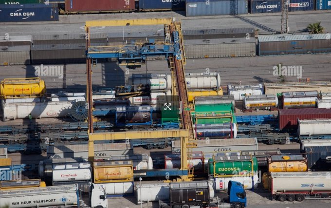 An aerial view of a busy shipping yard filled with stacked containers from companies like CMA CGM, GEODIS, Eurogate, and Van Heur Transport, alongside tank containers from Kampag and Cochland, being moved by a multi-rail gantry crane. The image serves as a visual metaphor for $ZBCN, illustrating how the crane operates across multiple parallel rails (blockchains/payment networks) to transfer cargo (money) between them, just as an overledger protocol functions above and across multiple underlying rails rather than being a single rail itself.
