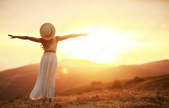 A woman in a white dress and sun hat stands on a hilltop with arms outstretched, facing a glowing sunset over distant mountains.