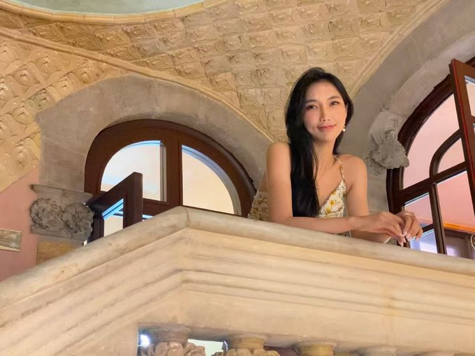 The image features a woman posing for a photo while leaning against a stone wall or balcony railing. She has her arms resting on the railing as she smiles at the camera, giving off a relaxed and confident vibe. The background of the image includes an archway with a window, adding to the overall aesthetic of the scene.