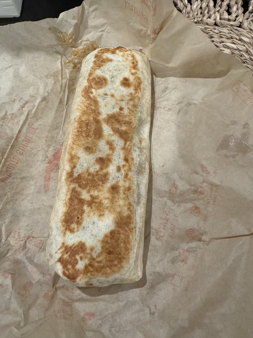 A golden-brown grilled burrito wrapped in Taco Bell paper packaging rests on crumpled wrapper, reflecting the post's humorous take on "white girl" trends with a focus on Taco Bell eating habits.