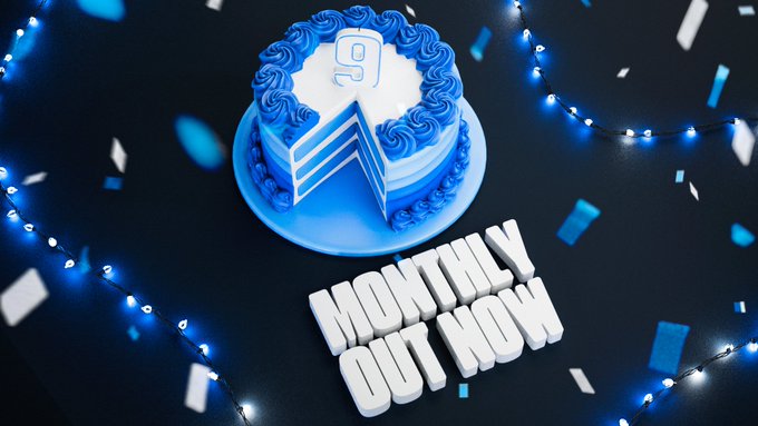 A blue and white birthday-style cake with a slice removed, decorated with a "9" candle on top, sits beside bold text reading "MONTHLY OUT NOW" on a dark surface adorned with blue string lights and scattered confetti. The image directly supports the post celebrating the 9th month of "Stake Monthly," creatively using a "9" cake to mark the milestone while the "MONTHLY OUT NOW" text reinforces the announcement.