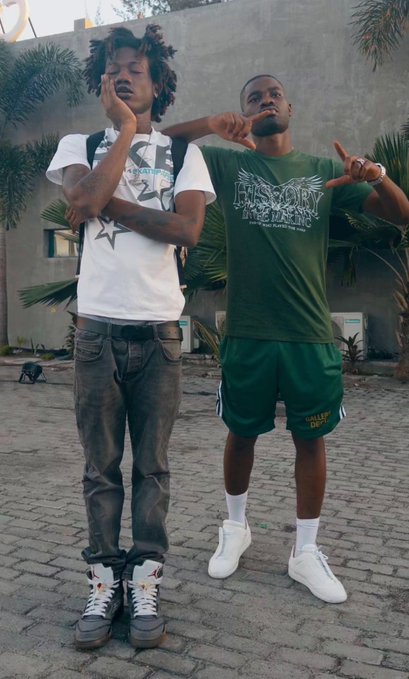 Two young men posing on a paved area in front of a building; one wears a white t-shirt, black jeans, and Air Jordans, while the other wears a green "JESUS CHRIST" t-shirt, green "COLLEGE DEPT" shorts, white socks, and sneakers.