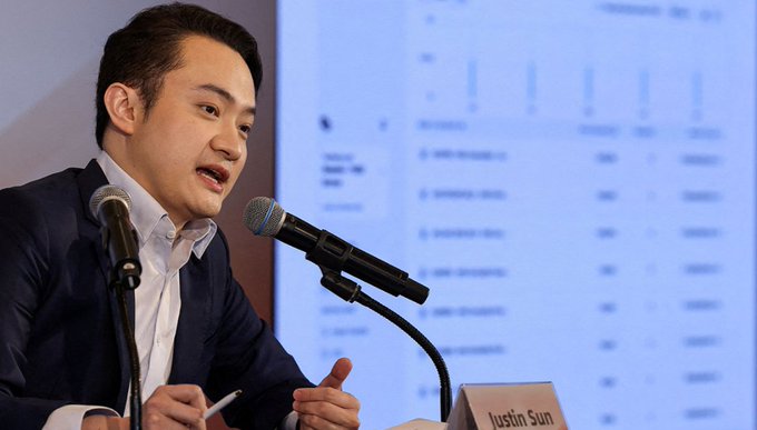 Justin Sun, founder of TRON, speaking into a microphone at a formal event or panel.