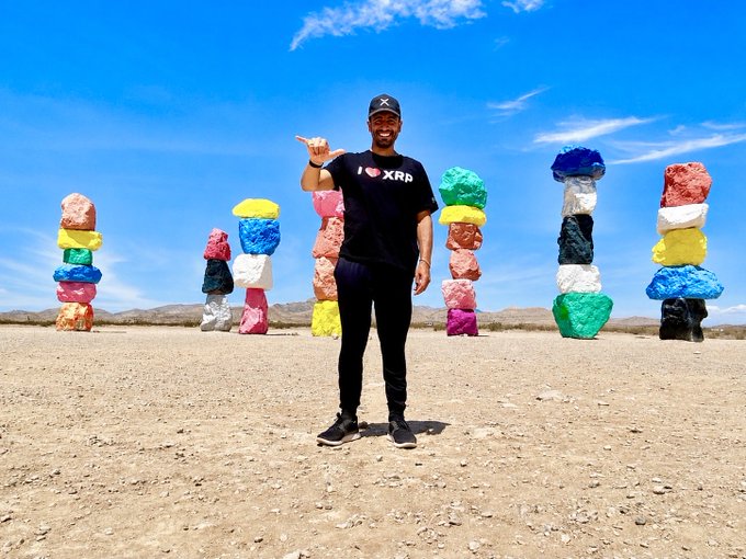 A man wearing a black 'I ❤️ XRP' t-shirt and cap points at the camera while standing in a desert landscape in front of colorful stacked rock sculptures.