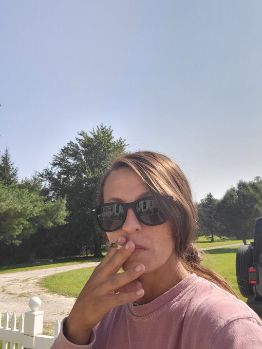 A woman with long brown hair, wearing black sunglasses and a pink sweatshirt, smokes a cigarette outdoors on a sunny day with a white picket fence and trees in the background. The image ties to the "Ladies of BAYC" post by showcasing a casual, candid moment of a woman enjoying a smoke break, fitting the laid-back theme of the Bored Ape Yacht Club community's female members.