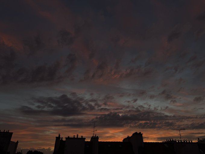 A dramatic sunset with deep crimson and orange clouds stretches above a silhouetted skyline of rooftops and chimneys, capturing the evening view from the perspective of someone taking a break from studying. The warm, fiery hues fading into the twilight sky evoke the late-night exhaustion of students pausing to appreciate the brief beauty before returning to their books.