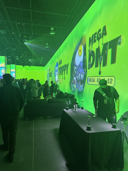 A brightly lit event space bathed in green light features large neon green banners reading 'MEGA DMT' with cartoon graphics, while people mingle near a staff member in a black cap.