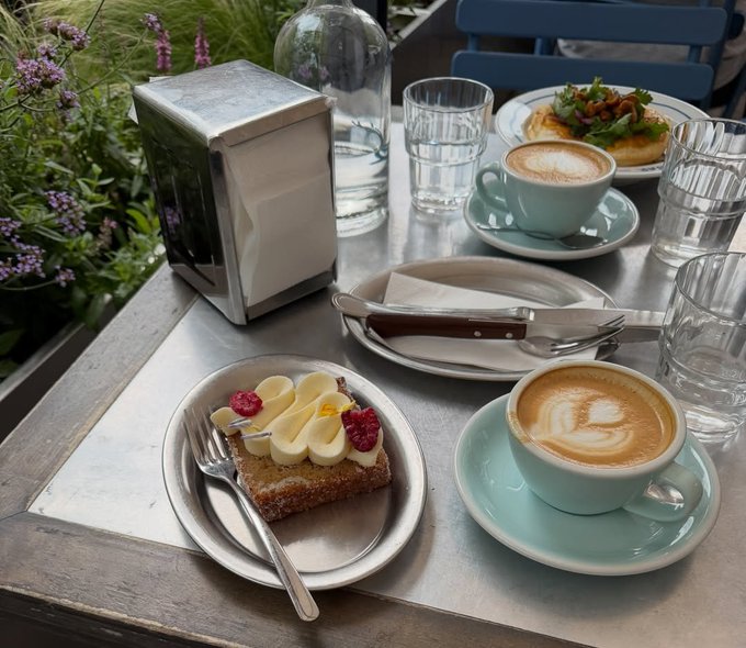 An outdoor cafe table with two lattes, a piece of toast topped with banana slices and raspberries, a savory dish with salad, water glasses, and a napkin dispenser.