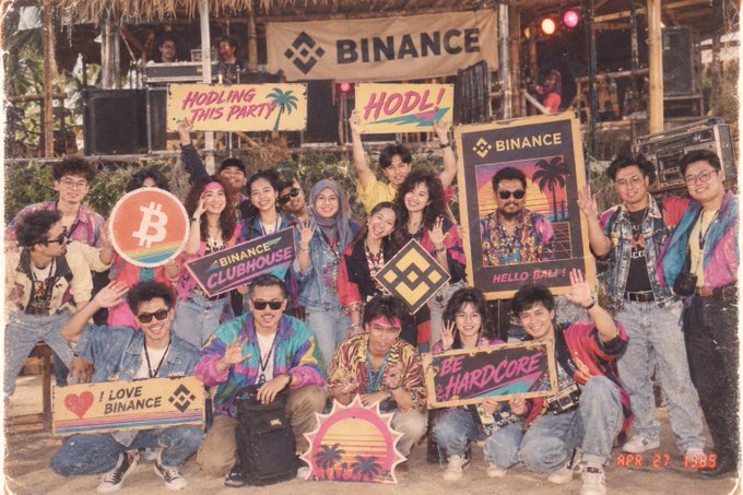 A group of people posing at a 'Binance' event with '80s' themed signs reading 'HODLING THIS PARTY', 'HODL!', 'HELLO SBN!', 'BE HARDCORE', and 'I LOVE BINANCE'.