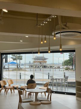 A person sits alone inside a bright, modern café with floor-to-ceiling windows, gazing out at the Yeongildae Pavilion in Pohang. The scene captures the quiet atmosphere of "Bitcoin CafeCafe Jomedical," Pohang's first Bitcoin-accepting café located on the basement level of Jomedical.