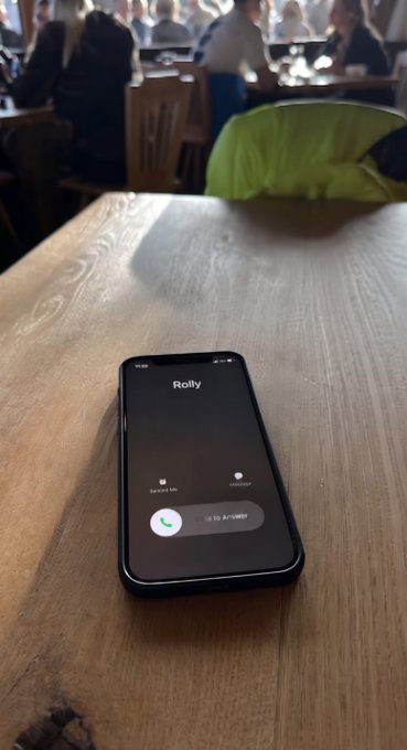 An incoming call from "Rolly" lights up a smartphone screen resting on a wooden table in a busy venue, with options to "Remind Me" or "Message." The post "Wins are calling. Wyd?" cleverly plays on the literal incoming call from a contact named Rolly, suggesting an exciting opportunity or victory is trying to reach the person—and asking if they're available to answer it.