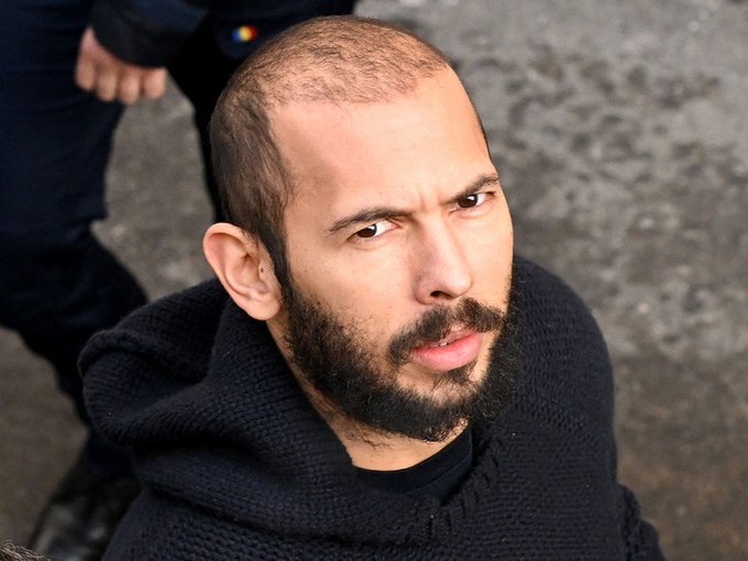 A man with a short buzz cut and dark beard looks up at the camera with an intense expression, wearing a black knit hooded sweater against a concrete background.