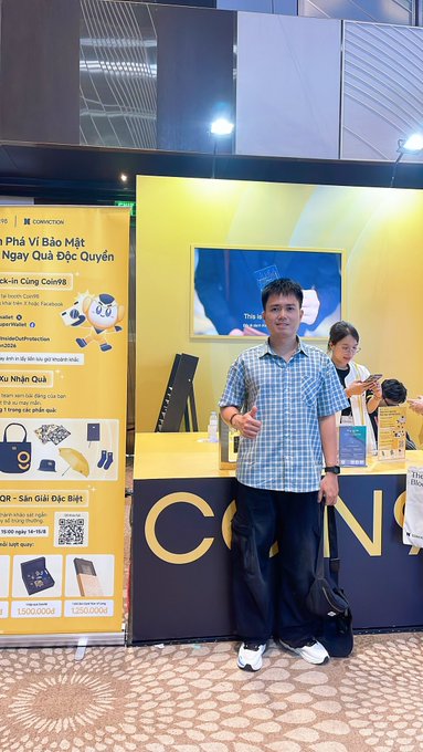 A photograph of a man in a blue checkered shirt standing and giving a thumbs-up in front of a yellow 'COIN98' promotional booth, with a promotional banner about copyright protection visible to the left.