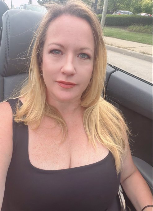 A selfie of a woman with blonde hair and blue eyes wearing a black scoop-neck top, sitting in a convertible car.