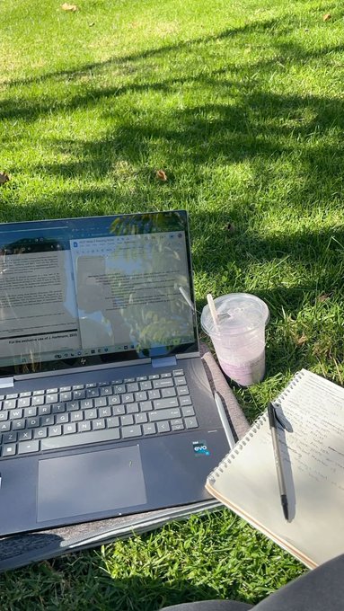 A laptop with an Intel Core sticker, an iced drink, and an open spiral notebook with a pen are arranged on green grass for outdoor work or study.