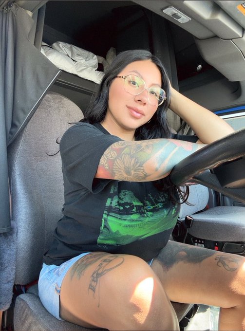 A young woman with glasses, long dark hair, and visible tattoos on her arm and thigh sits in the driver's seat of a truck, wearing a black graphic t-shirt and light denim shorts, with her hand resting behind her head and a pillow visible in the sleeper cab behind her.