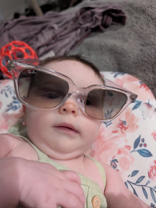 A baby wearing oversized clear-framed sunglasses lies on a floral-patterned surface, looking at the camera.