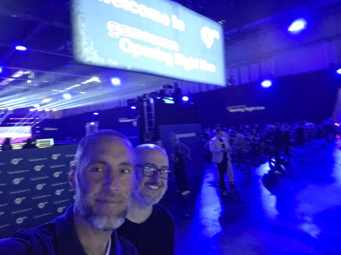 Two men take a selfie at a technology event, with a large screen above them displaying "Welcome to gameaccess" and "Opening Night Live," while a "Gaming Reaktor" booth is visible in the background, setting the scene for their attendance at the ONL (Opening Night Live) showcase.