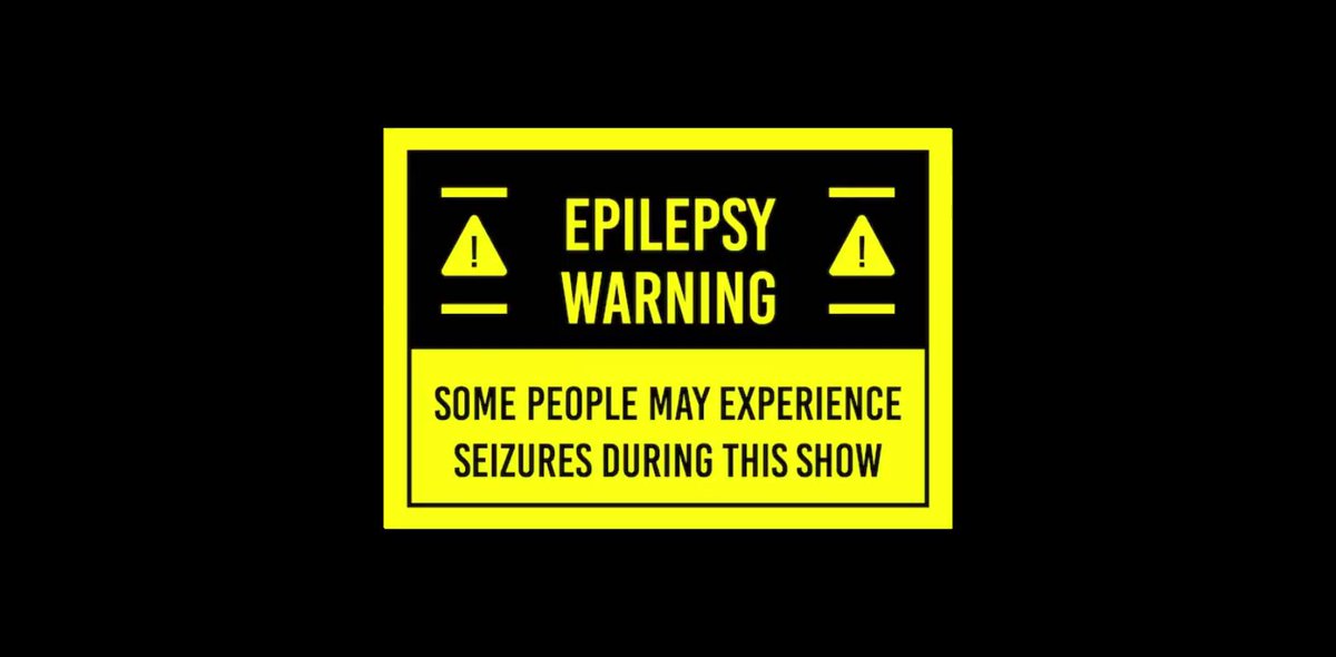 The image features a warning sign that reads "Epilepsy Warning" with yellow text against a black background, accompanied by a yellow border. The sign also includes two small red triangles with a white outline, which are often used as visual indicators of potential hazards or risks in an area.