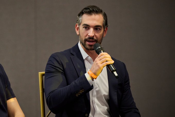 A man with a beard wearing a dark suit jacket over a white shirt, speaking into a microphone while seated on stage, wearing an orange wristband.