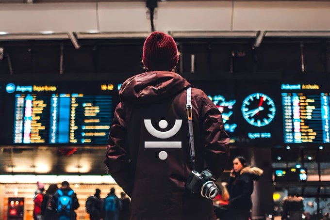 A person in a maroon hooded jacket stands in front of illuminated flight information departure boards and an analog clock in an airport terminal, with a camera hanging from their shoulder.