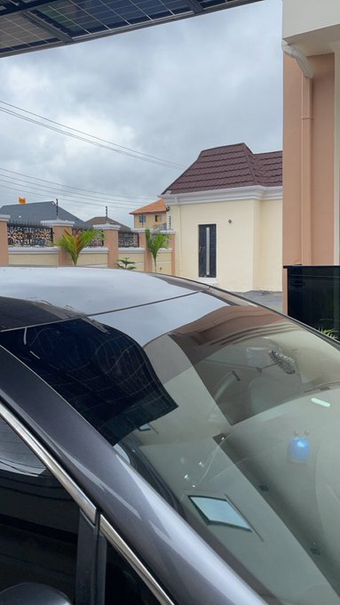 A view from under a carport looking out at a residential street with houses, a cloudy sky, and the glossy roof and windshield of a parked dark car.