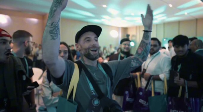 The image shows a man with tattoos standing up and raising his arms in the air while holding a camera bag. He appears to be at a social event or gathering where he has won a new camera as a prize. The surrounding people are also present, indicating that this is an event of interest for many attendees.