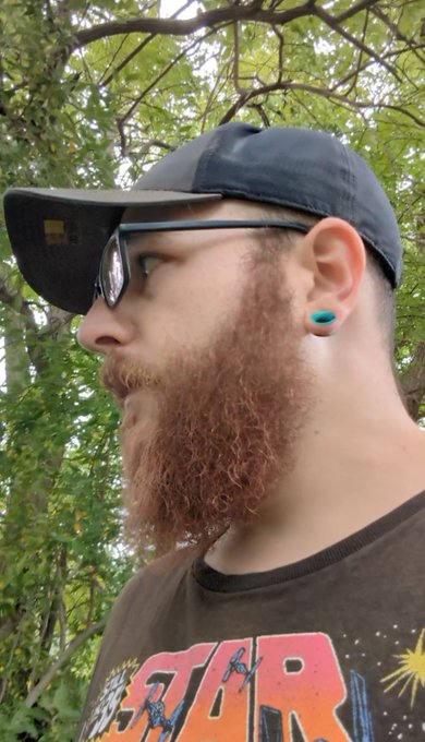 A bearded man in a black cap and glasses, wearing a "Star Wars" t-shirt and a turquoise ear gauge, poses for a selfie in a wooded outdoor setting, matching the post's "touched grass" theme and its running $ADA $SNEK price-tracking series.