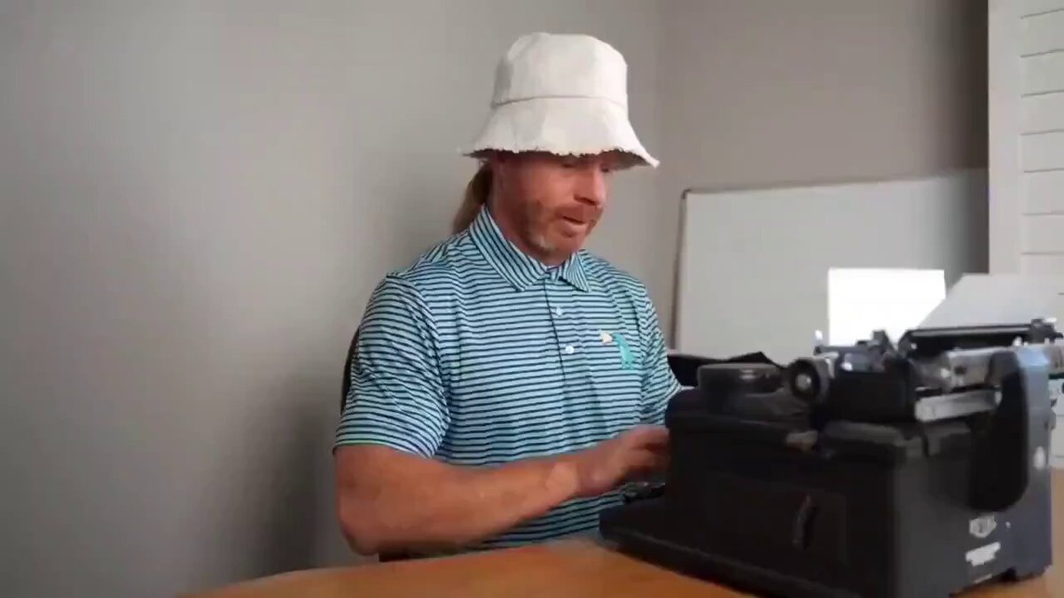 The image features a man wearing a white bucket hat sitting at a desk with an old-fashioned typewriter, typing on it. The typewriter has a black cover and appears to be quite large compared to the man's size. The background of the image is a plain gray wall, which contrasts with the vibrant colors of the typewriter and the man's attire.