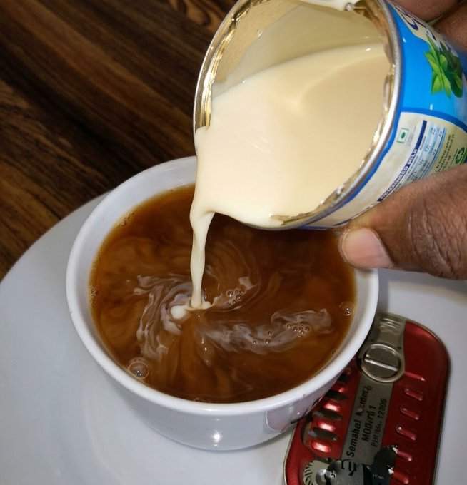 A close-up shot of cream or condensed milk being poured from a tin can into a cup of dark tea or coffee, sitting on a white saucer next to a red multi-tool with "Semahal M1004-01" visible.