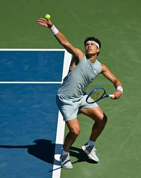 Tennis player Zheng Qinwen serving on a blue hard court, tossing a yellow tennis ball while holding a racket mid-motion.