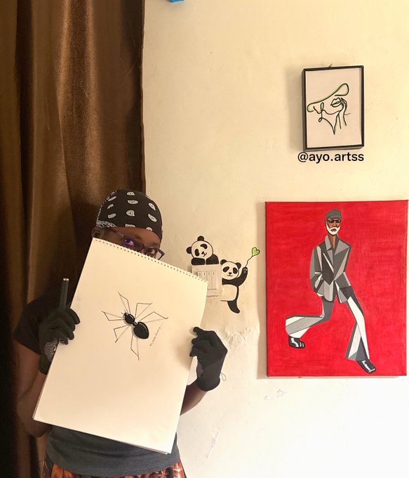A person wearing a black bandana, glasses, and black gloves holds up a sketchbook displaying a drawing of a black spider, with artwork including a red canvas featuring a man in a suit visible on the wall behind them, tagged "@ayo.artss."