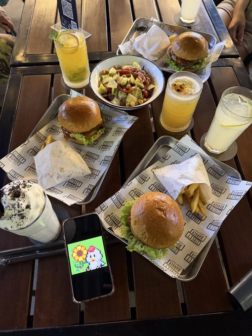 A wooden table with three burgers, fries, salads, and multiple drinks, with a smartphone displaying a pixel art chicken.