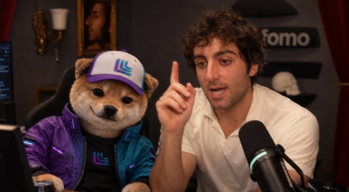 A man in a white shirt gestures with his index finger raised while speaking into a microphone, sitting next to a Shiba Inu mascot wearing a purple jacket and white cap with a 'W' logo.