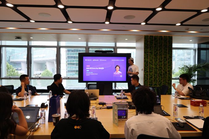 A group of people gathered in a conference room with a presentation screen displaying "Injective Demo Day" with a speaker presenting to attendees seated around a table. The image relates to the post by showcasing Injective's commitment to supporting early-stage builders through events like Demo Day, which provides the guidance, distribution, and ecosystem support mentioned in the caption.