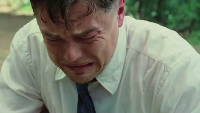 A close-up shot of a man in a white shirt and dark tie with a pained, crying expression, appearing to be outdoors with greenery in the background.